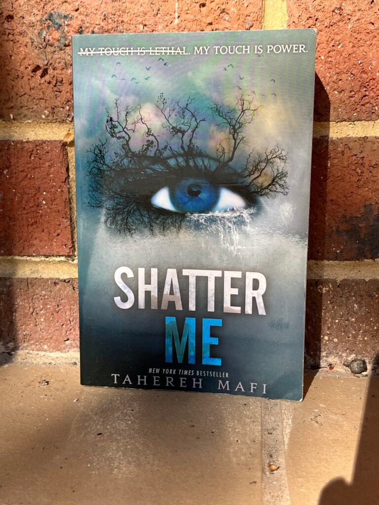 The image is over the cover of a book. The t title is 'Shatter Me'. The cover has an image of a blue eye with trees growing out of the eye and cloudy skies as the background. The book is photographed in front of a brick wall.